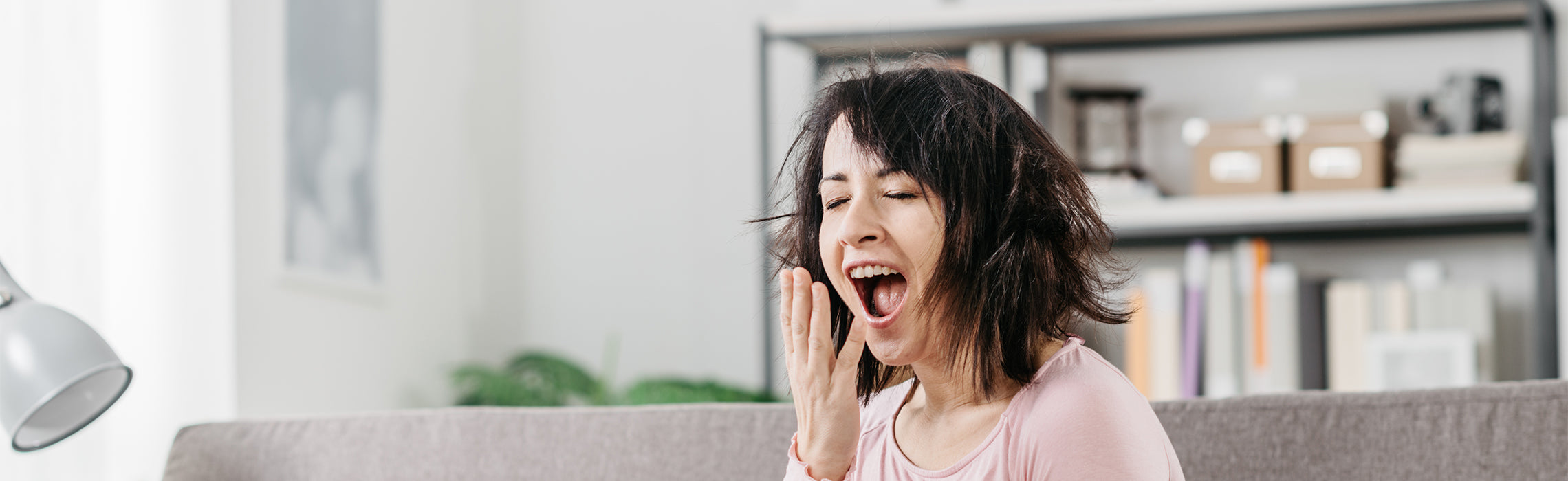 Person sitting on a couch indoors with eyes closed and mouth open while covering the mouth with one hand mid-yawn, with a lamp, plant, and shelving in the background, representing fatigue, tiredness, and lack of sleep.