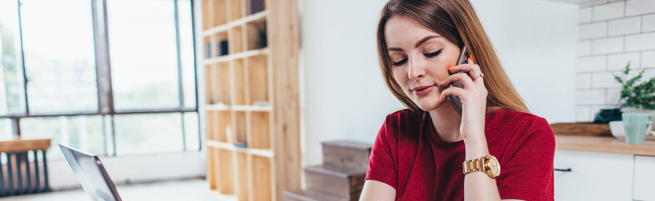Person in a red shirt sitting at a desk, talking on a smartphone beside an open laptop in a bright home office with shelves and a window in the background. No visible text or branding.