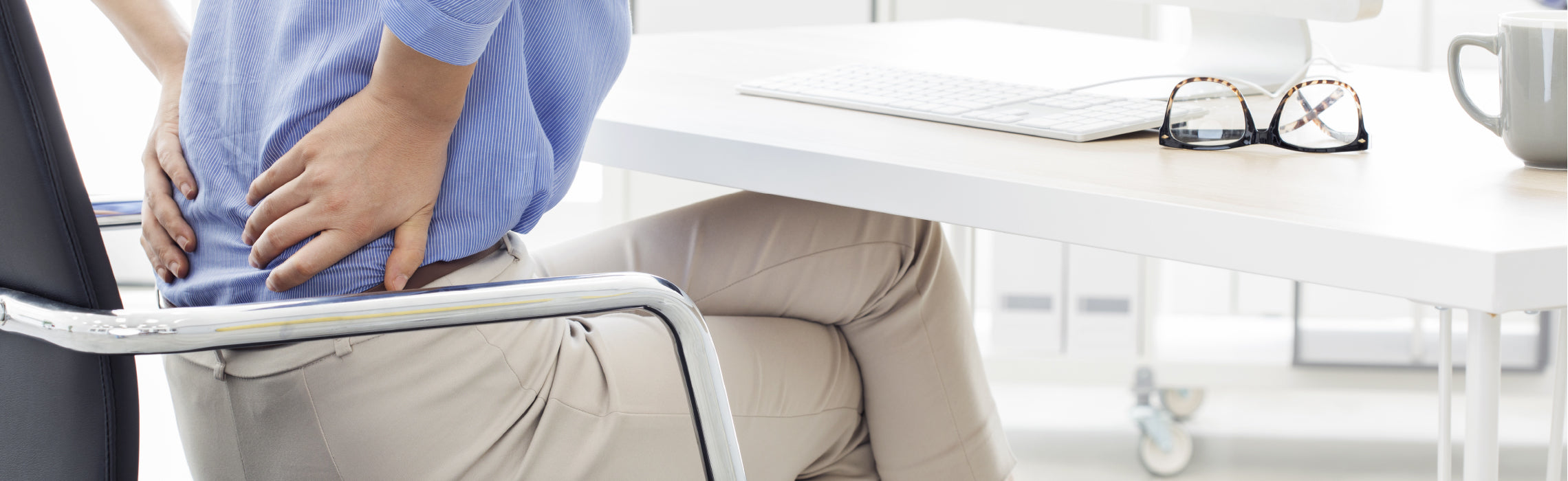 Person seated at an office desk holding the lower back with one hand while leaning in a chair, with a laptop, glasses, and coffee mug nearby, representing lower back pain, poor posture, and workplace strain.