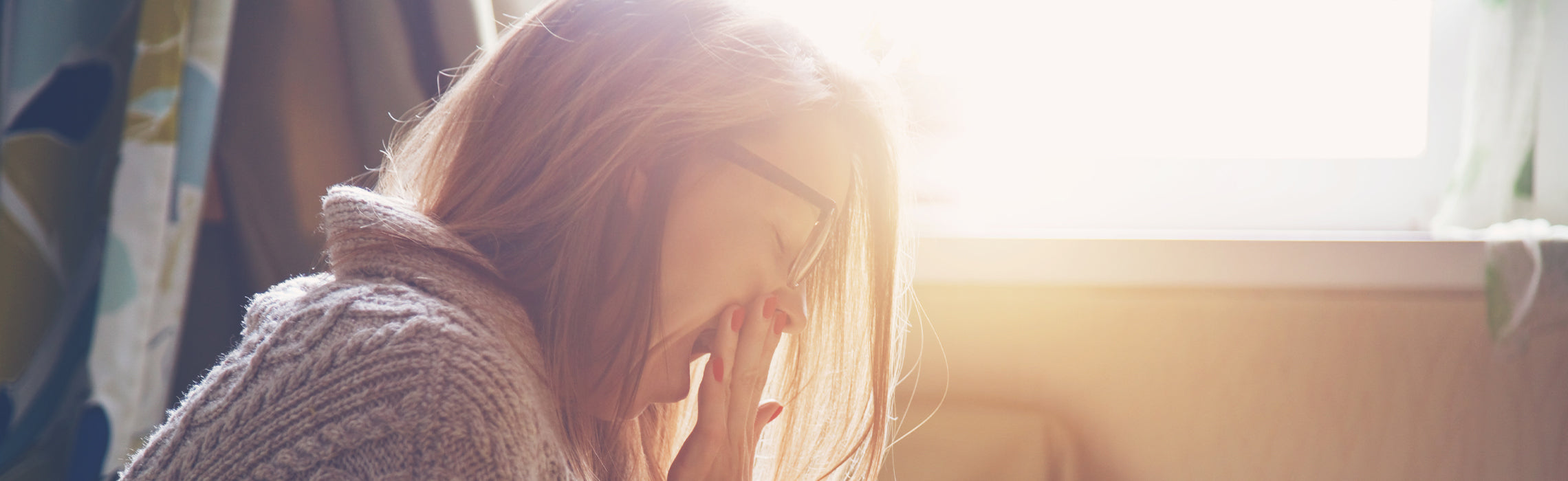 Woman sitting indoors in soft morning light, holding her forehead and appearing tired or stressed.