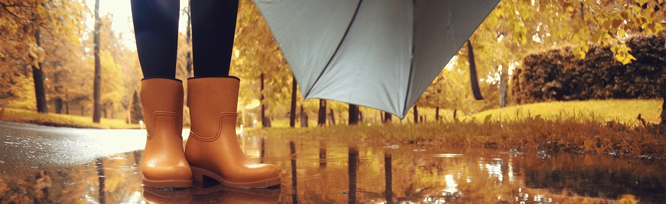 Person standing on a wet path wearing tan rain boots and holding an umbrella, with autumn trees and fallen leaves reflected in puddles, representing rainy weather, fall season, and outdoor activity.
