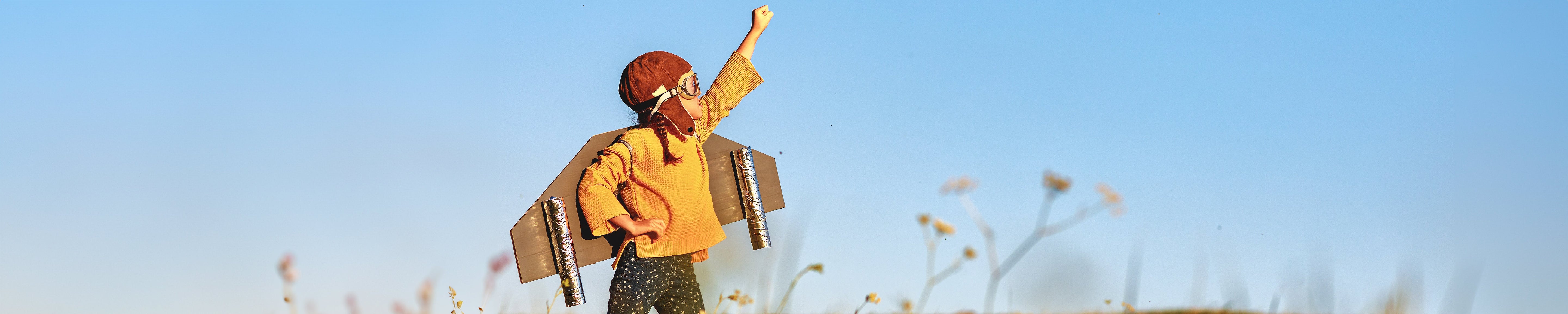 Child wearing a cardboard airplane costume and aviator goggles stands in a field with one arm raised toward the sky
