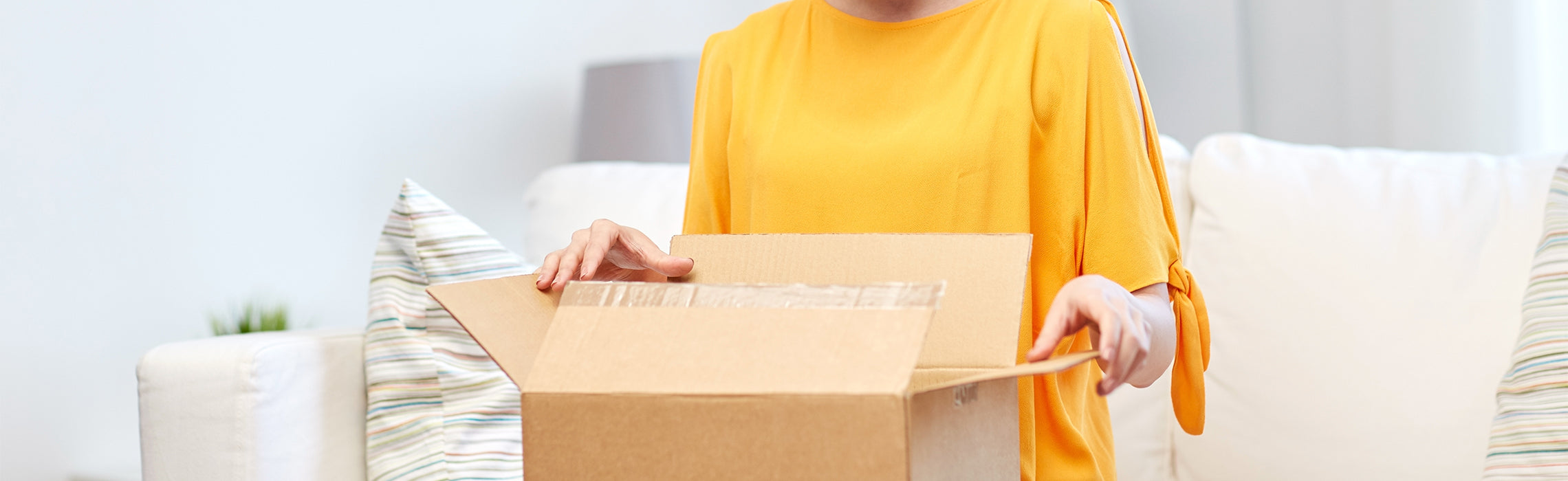 Person in a bright living room opening a cardboard box while seated on a couch, representing online shopping delivery, unboxing, and home retail experience.