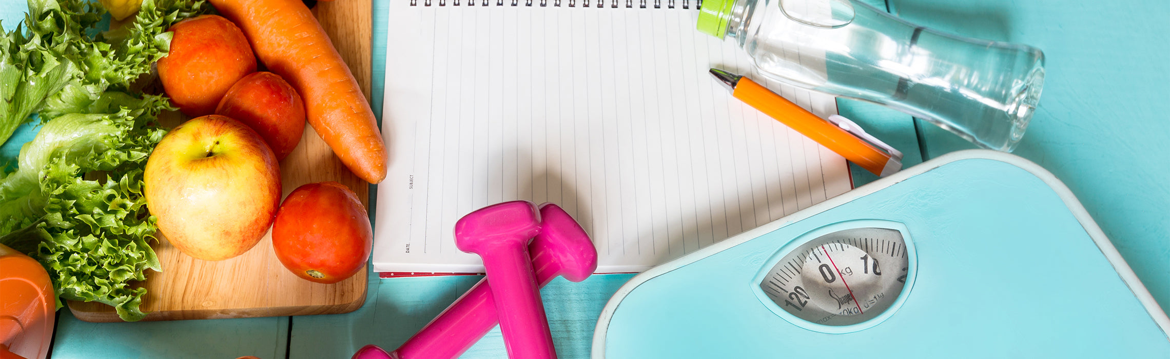Flat lay of fresh vegetables including lettuce, carrots, and apples beside a blank lined notebook, pink dumbbells, a water bottle, pen, and a bathroom scale, representing meal planning, fitness goals, weight loss, and healthy lifestyle tracking.