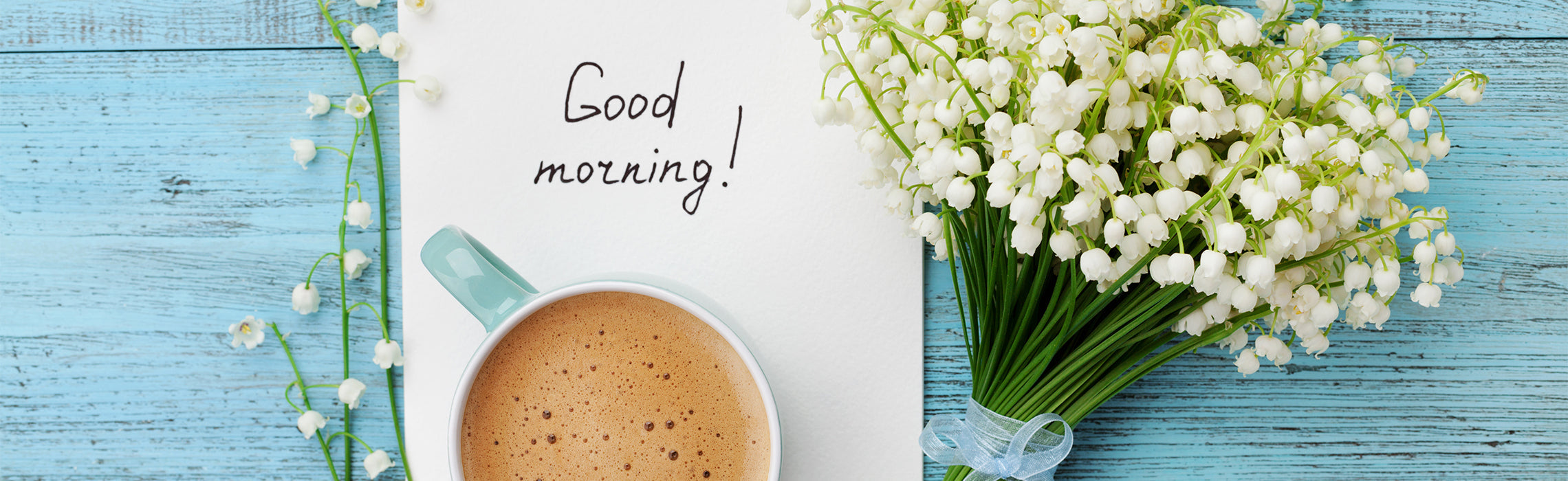 Flat lay of a coffee cup, notebook with handwritten text “Good morning!”, pen, and white flowers on a blue wooden surface.
