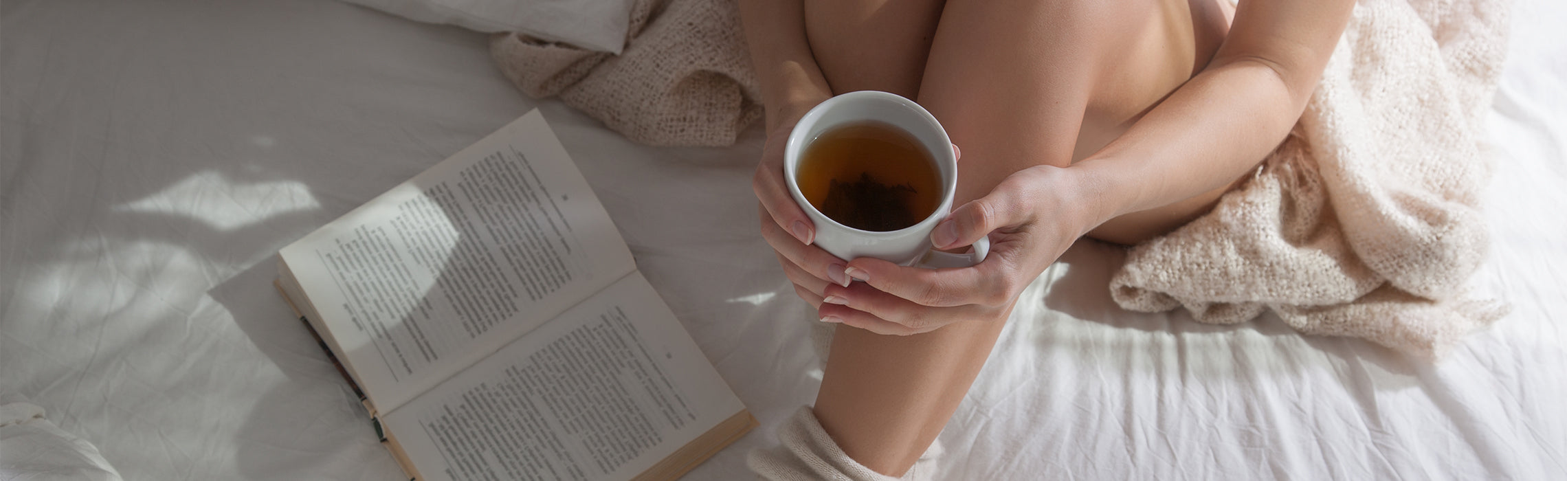 Person relaxing in bed while holding a cup of tea beside an open book, creating a calm and cozy self-care scene.