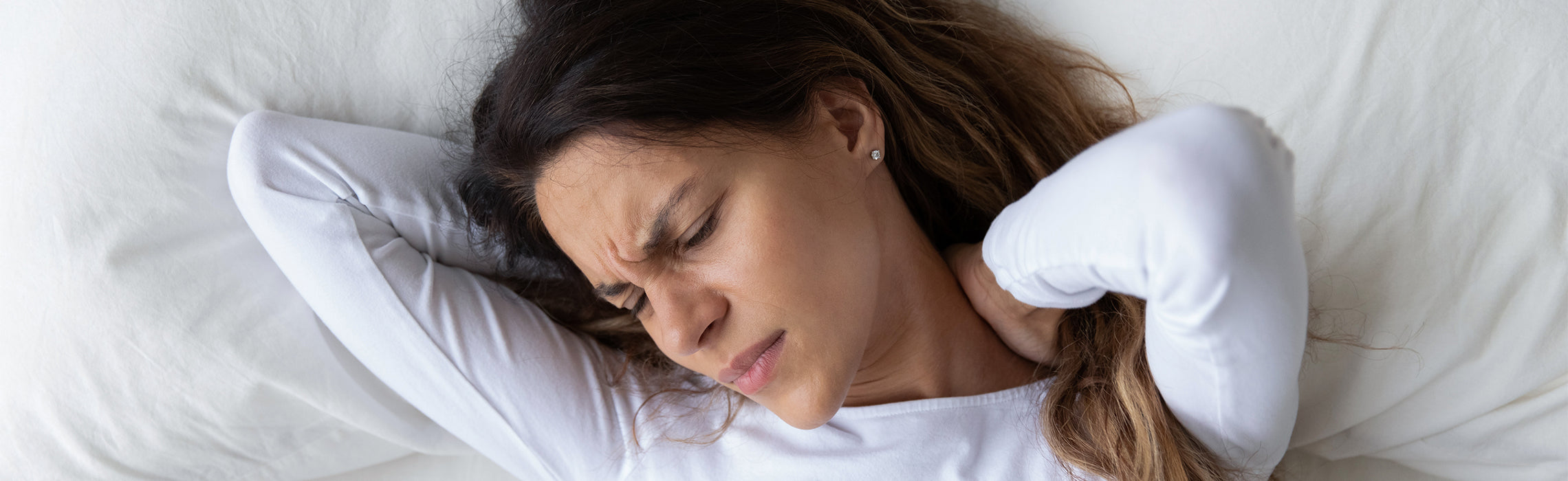 Woman lying in bed with hands behind her neck, appearing uncomfortable from neck pain or stiffness while resting on white bedding.