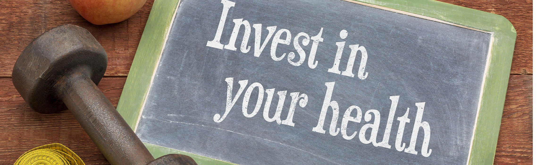 Chalkboard sign reading “Invest in Your health” placed beside a dumbbell and fresh fruit