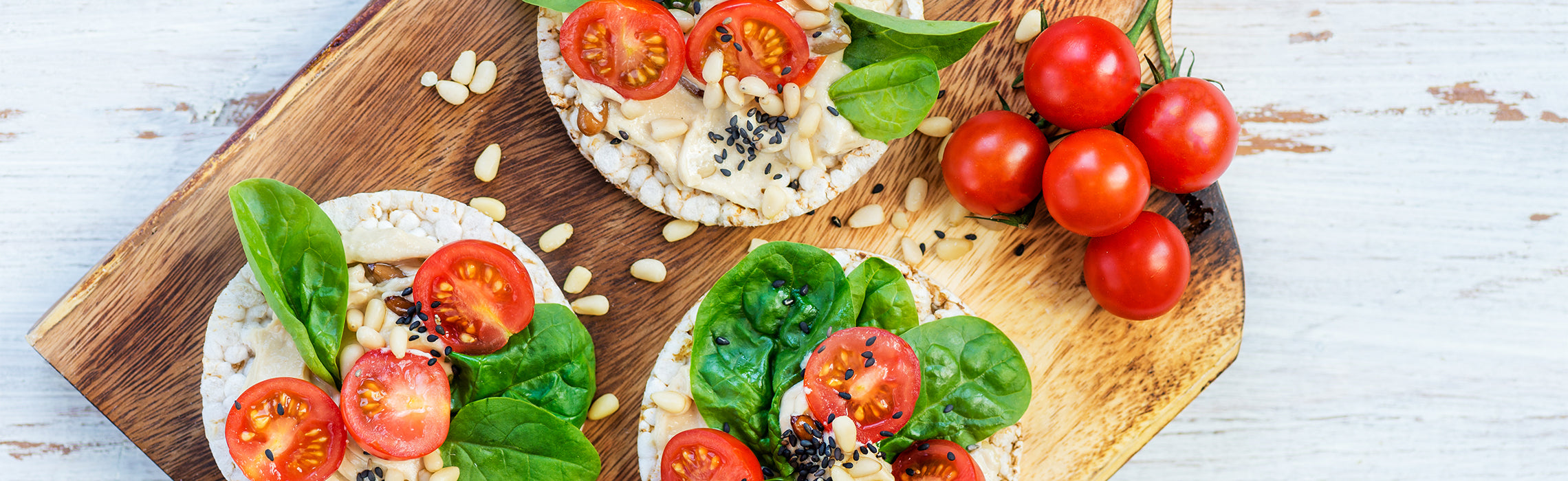 Top-down view of rice cakes topped with hummus, cherry tomatoes, spinach leaves, seeds, and pine nuts on a wooden board, with fresh vine tomatoes on the side, representing healthy snacks and balanced nutrition.