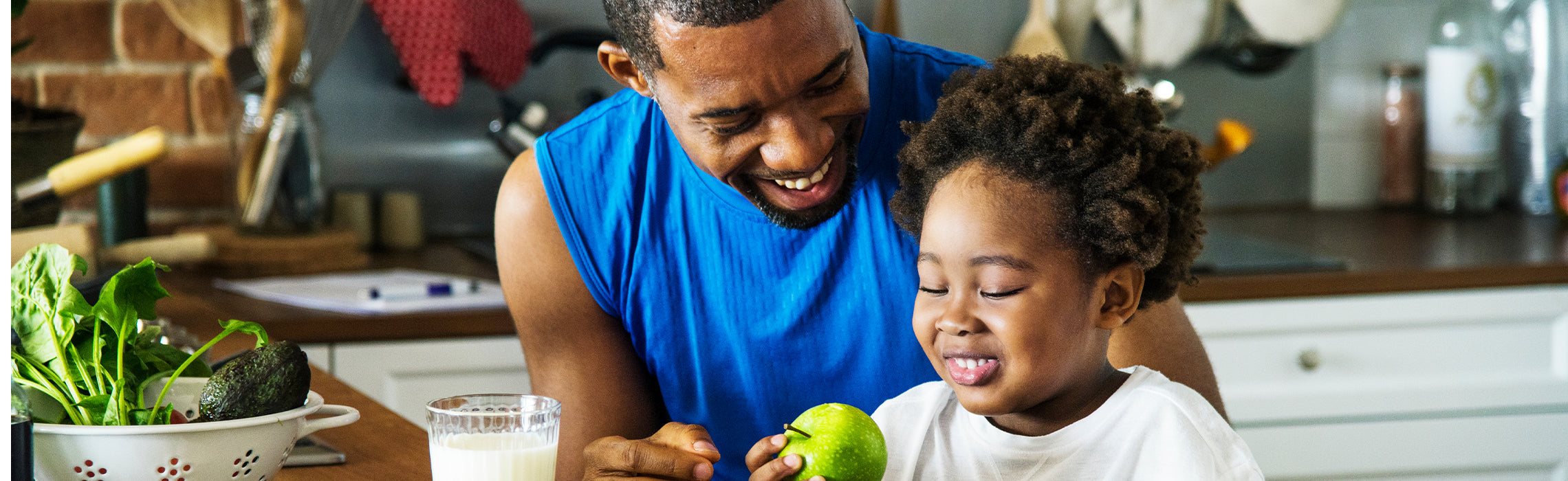 Adult and child at a kitchen table with fresh produce and a glass of milk, with the child holding a green apple while preparing food together, representing healthy eating, family nutrition, and wellness habits.