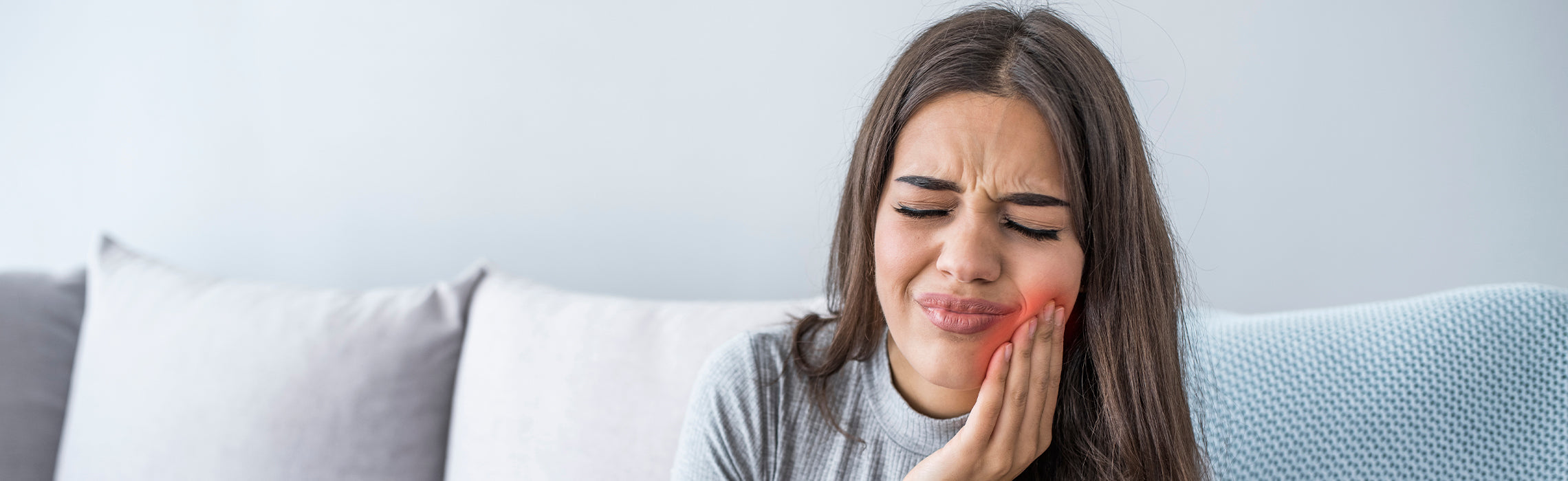 Person sitting on a couch pressing a hand against the jaw with a highlighted red cheek area, representing toothache, dental pain, or jaw discomfort in a home setting.