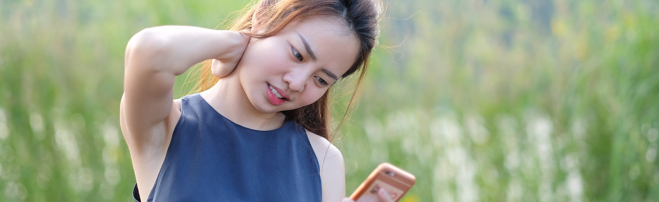 Person outdoors holding the back of the neck with one hand while looking at a smartphone, representing neck pain, muscle strain, and discomfort from mobile device use.