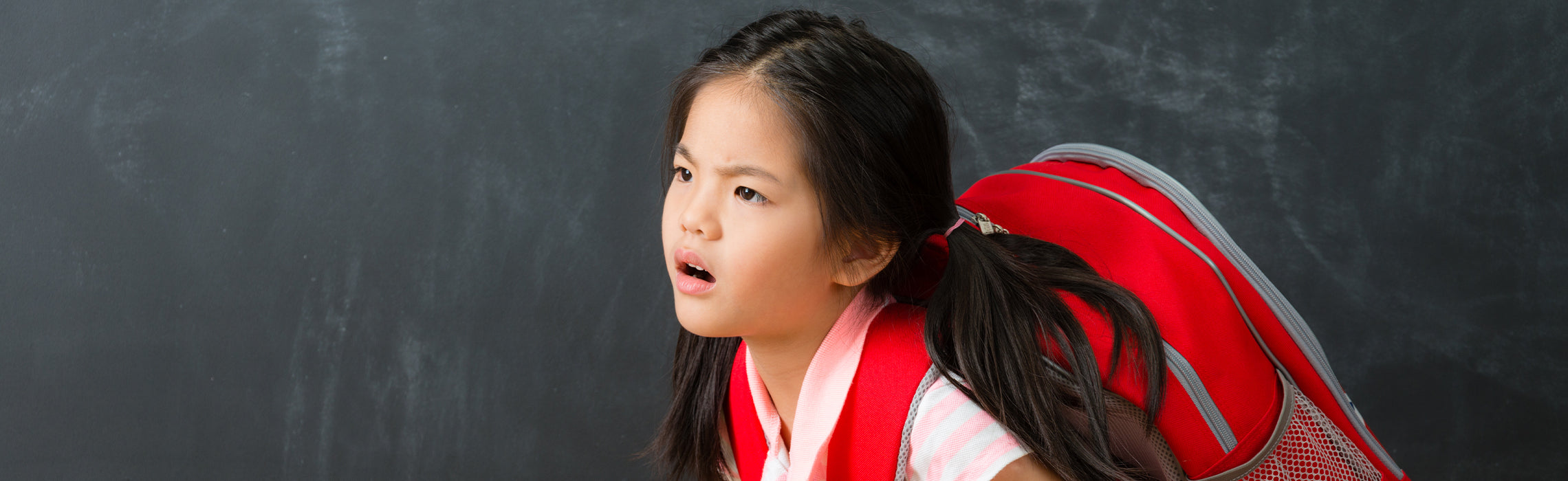 Child wearing a red backpack leaning forward in front of a chalkboard, representing school fatigue, heavy backpack strain, and student posture concerns.