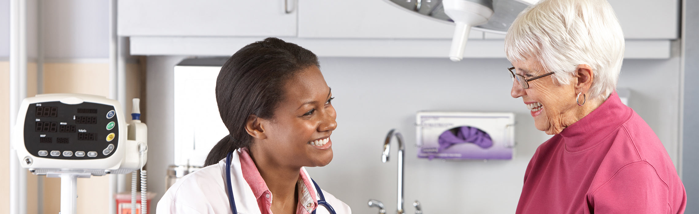 Healthcare professional in a clinical exam room speaking with an older adult, with medical equipment, cabinets, and a sink in the background, representing routine checkup, patient care, and medical consultation.