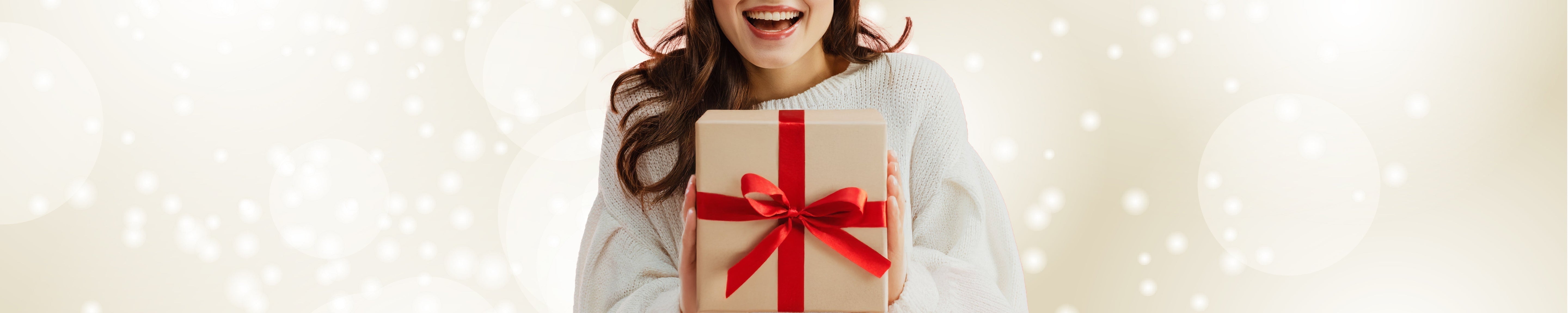 Close-up of a person holding a wrapped gift box with a red ribbon and bow, wearing a light sweater against a soft neutral background with bokeh lights, representing holiday gifting and celebration. No visible text.