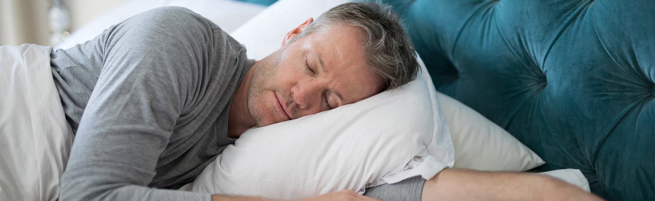 Person lying on a bed sleeping on a white pillow with gray bedding, next to a teal upholstered headboard, representing restful sleep, comfort, and proper sleep position in a calm bedroom setting.