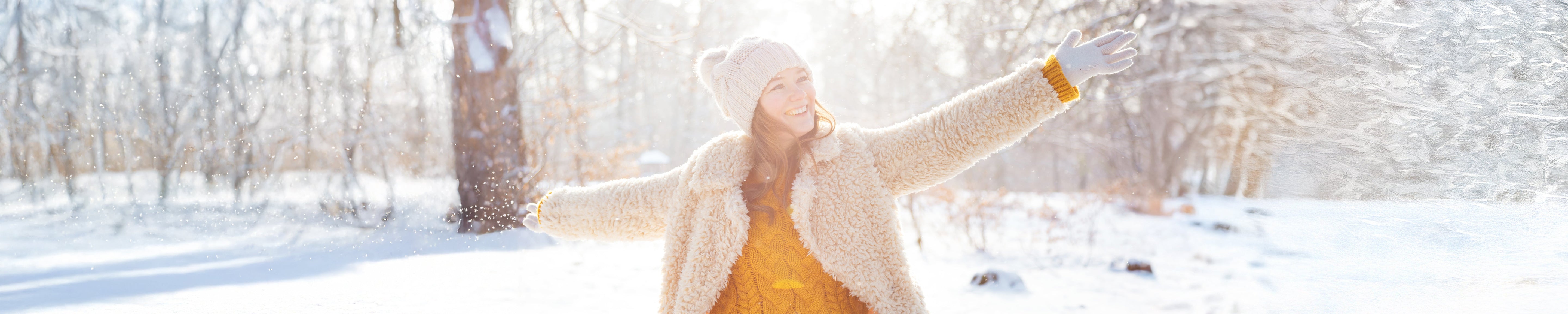 Person outdoors in a snowy park wearing a winter coat, knit hat, and gloves, standing with arms outstretched as snow falls in bright sunlight; trees and a frosty landscape in the background create a seasonal winter scene. No visible text.