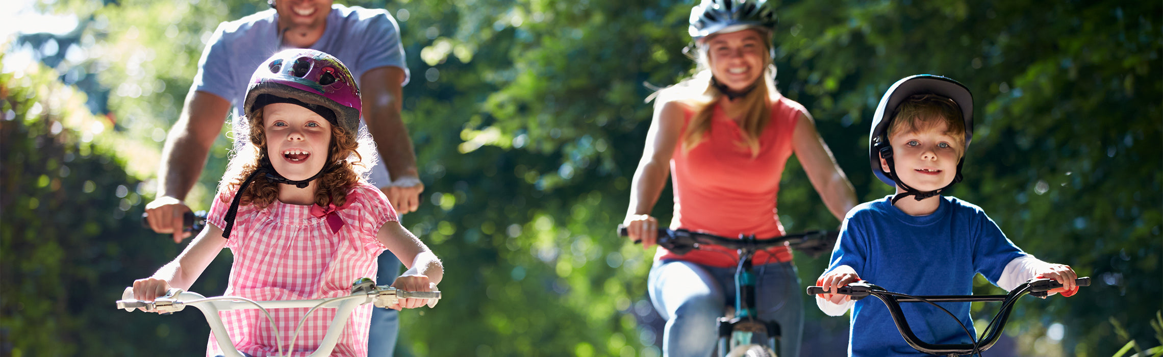 Family of four riding bicycles along a sunlit, tree‑lined path, with two children in helmets leading and two adults following, representing outdoor recreation, family fitness, and active lifestyle.