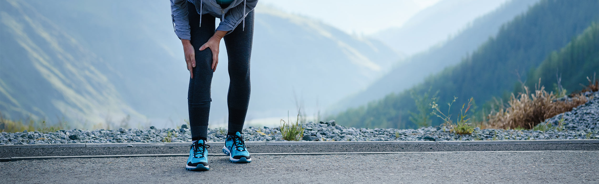Person outdoors on a roadside holding one knee with both hands, wearing blue running shoes and athletic clothing, with mountains in the background, representing knee pain, running injury, and outdoor fitness strain.