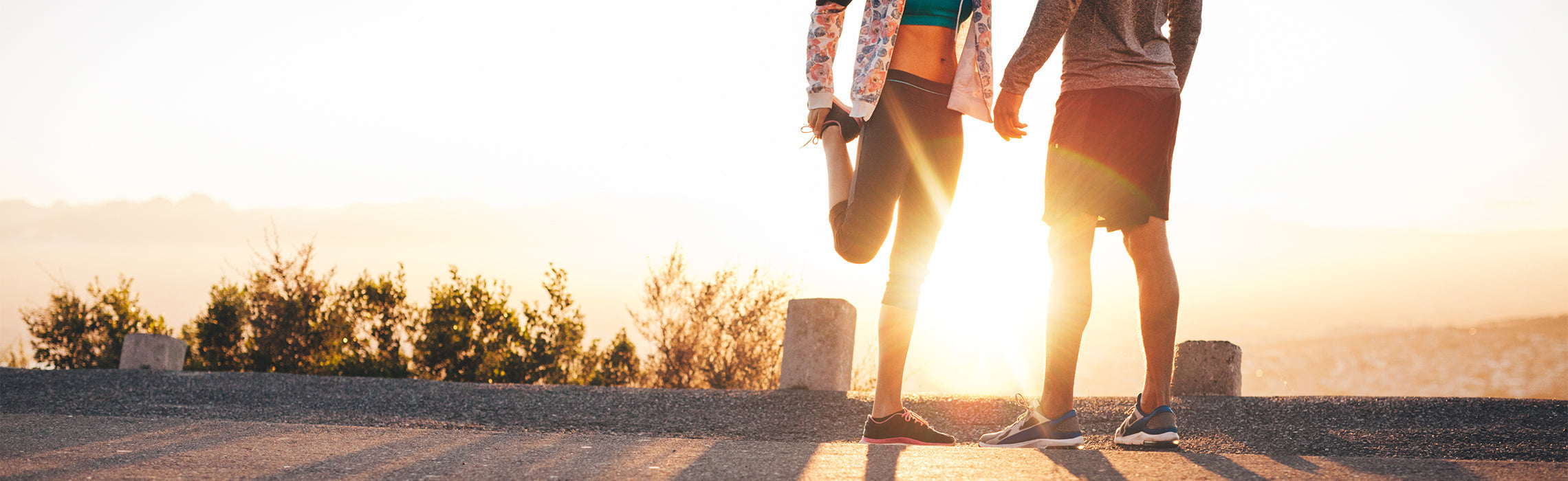 Woman stretching her leg beside another person outdoors at sunset on a paved path