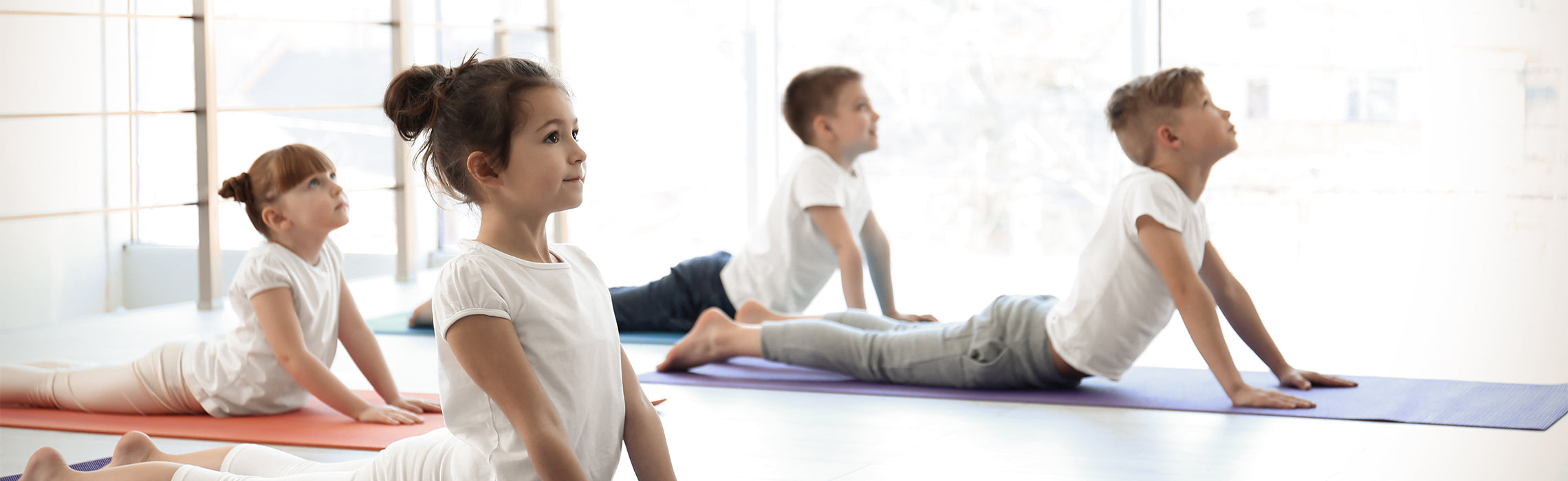Four children practicing yoga on mats in a bright studio, performing an upward stretch pose during a group fitness or wellness class. No visible text.