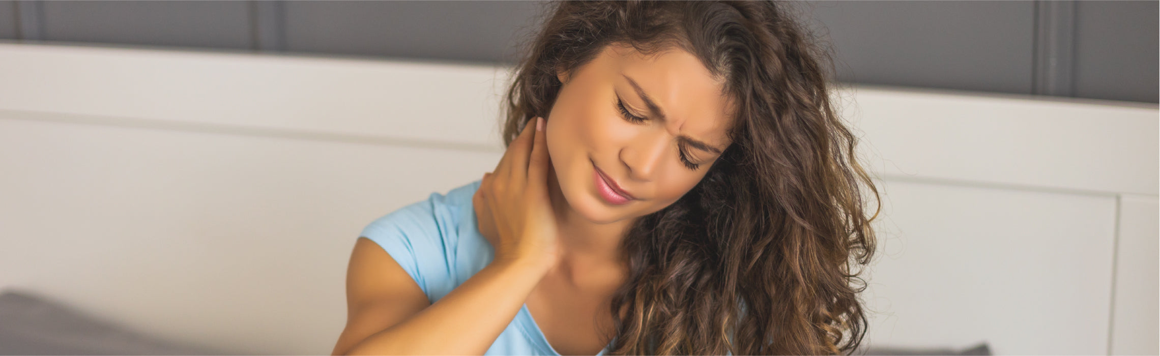 Close-up of a person indoors holding the side of the neck with one hand, indicating neck pain or muscle strain, with a neutral home setting in the background.