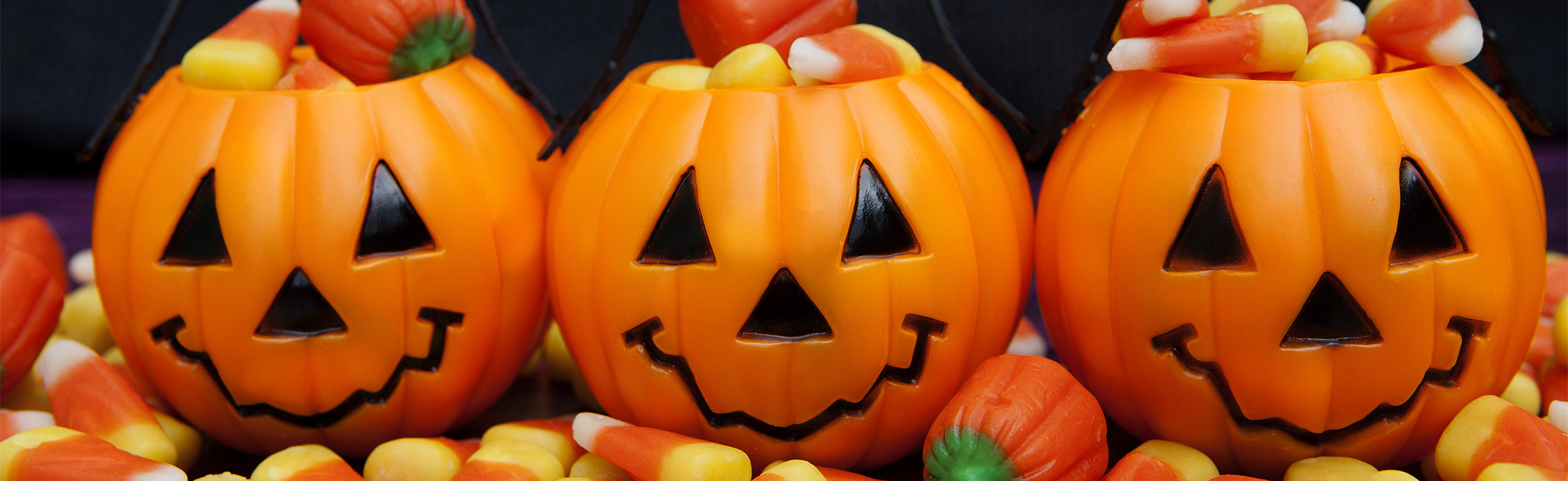 Three orange jack‑o’‑lantern candy containers filled with candy corn and fall treats, arranged close together against a dark background, representing Halloween decorations, seasonal sweets, and festive holiday décor.