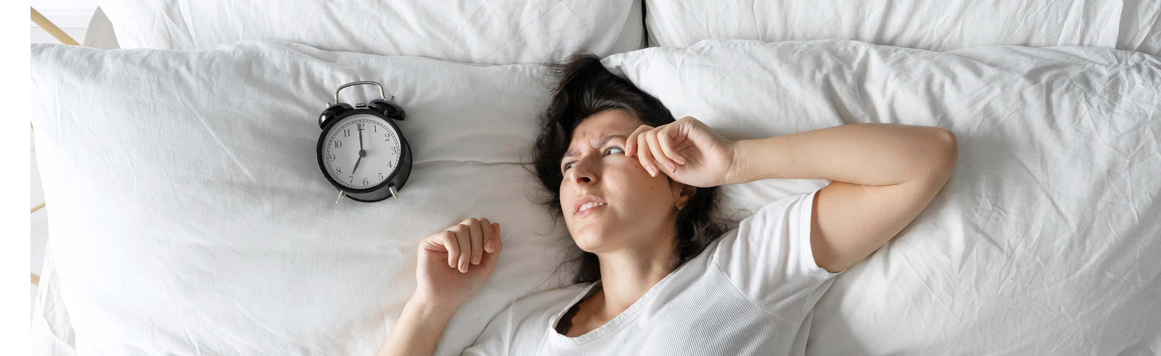 Woman lying awake in bed beside an alarm clock, appearing tired and restless on white bedding.