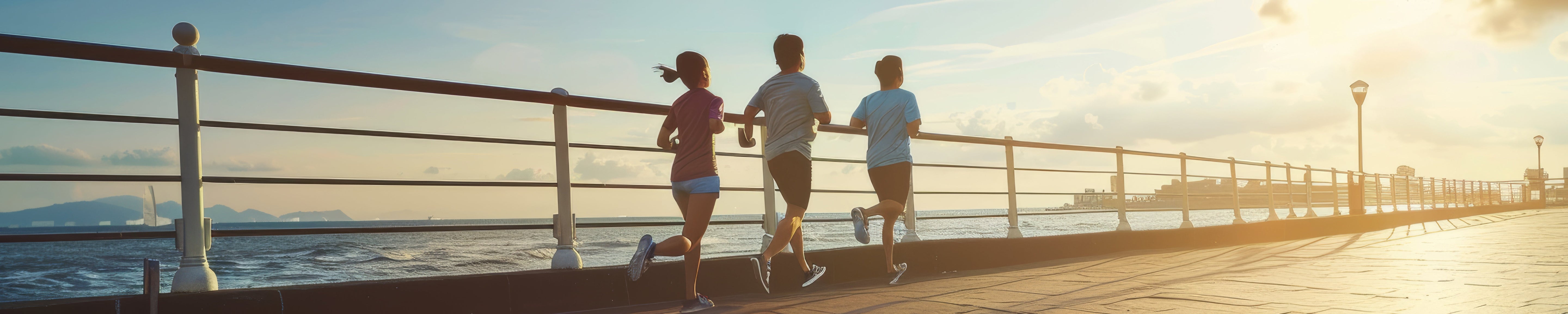 Three people jogging along a waterfront boardwalk at sunrise, running beside a metal railing with ocean views; warm sunlight and long shadows highlight an active outdoor fitness scene. No visible text.