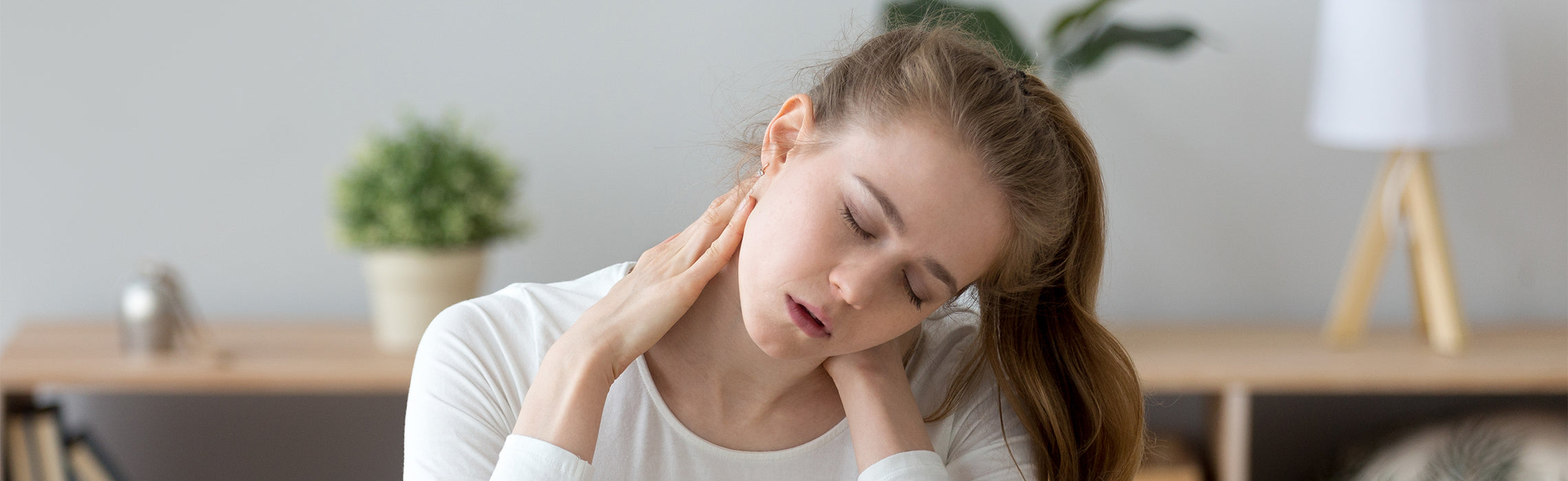 Person sitting indoors holding the neck with both hands and tilting the head downward, representing neck pain, muscle strain, and posture-related discomfort in a home setting.