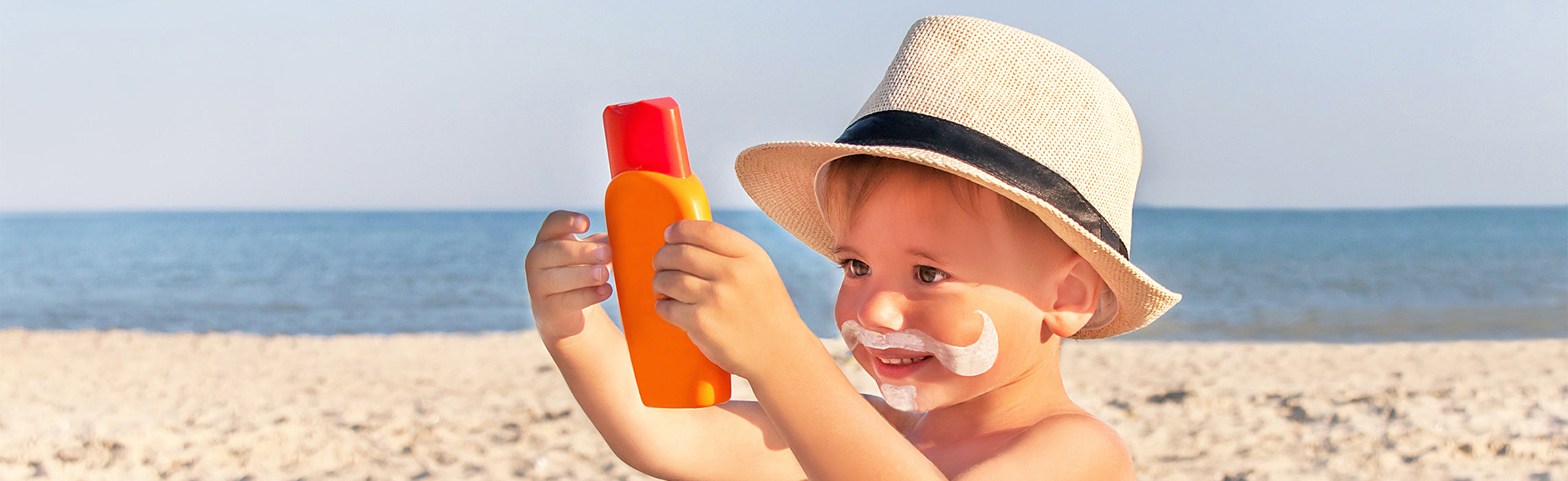 Young child wearing a sun hat and sunscreen on the beach while holding a bottle of sunblock.
