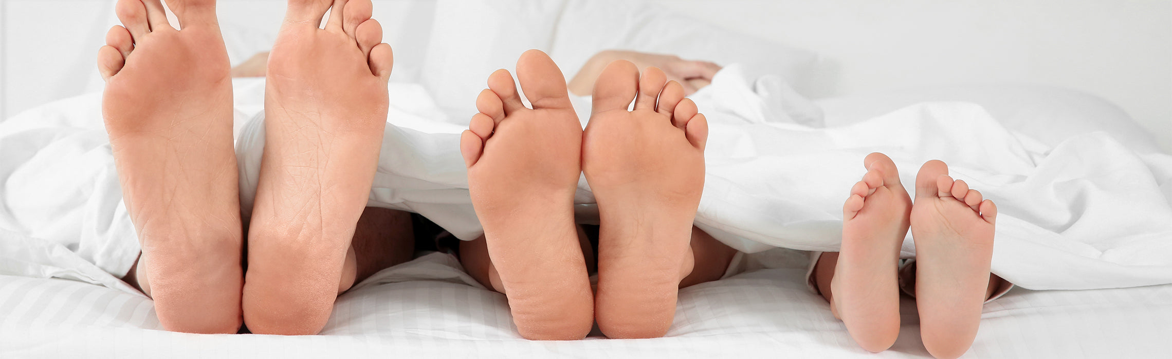 Close-up of three pairs of bare feet—two larger and one smaller—extending from under white bedding on a bed, representing family togetherness, comfort, and relaxed home life. No visible text.