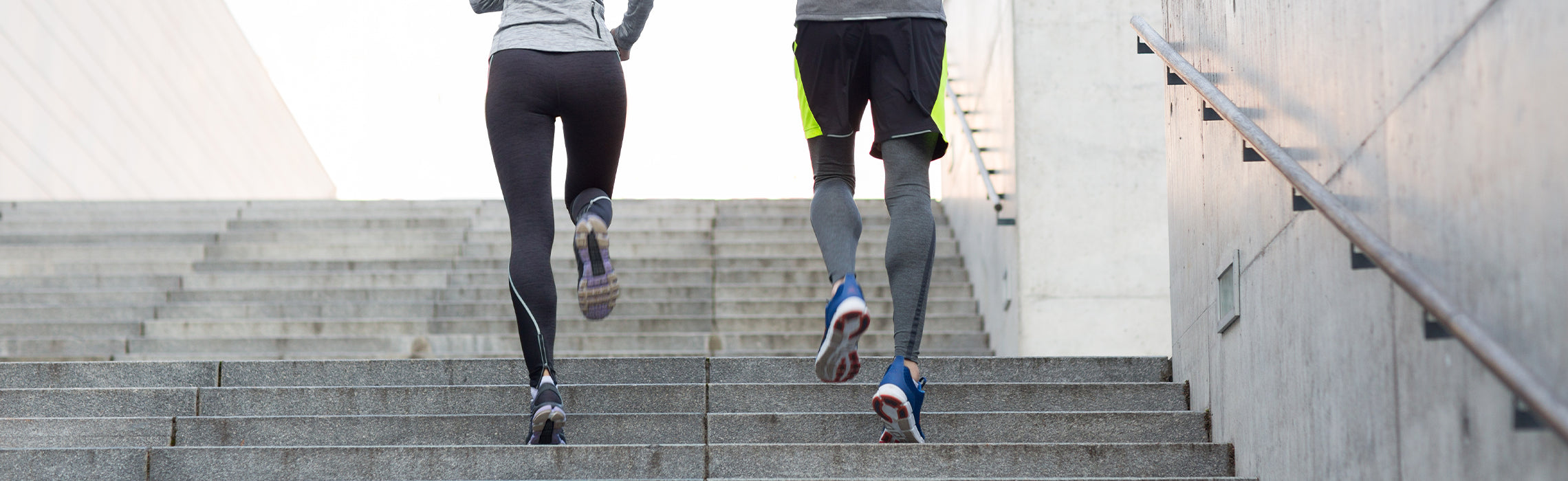 Two people in athletic wear running up outdoor concrete stairs, viewed from behind, highlighting fitness training and cardio exercise. No visible text or branding.