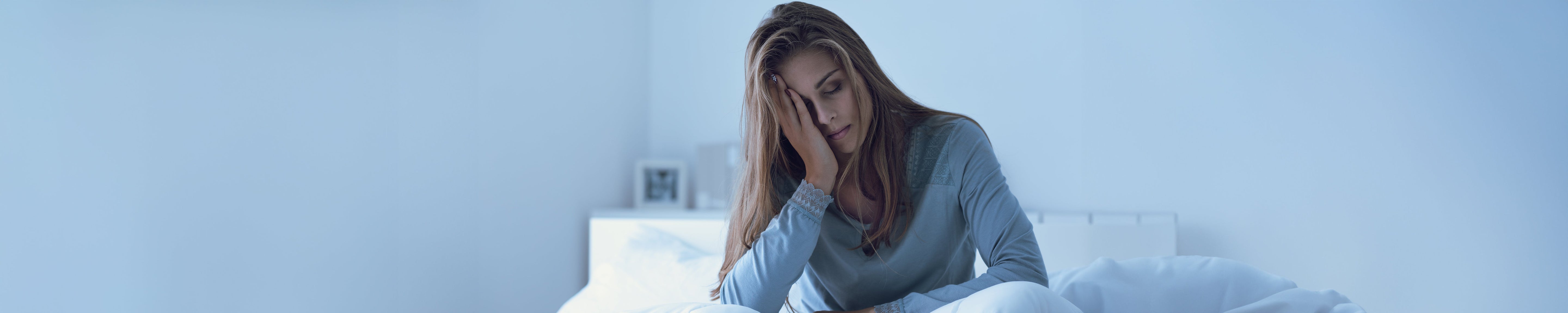 Person sitting upright in a bed at night, resting one hand on the forehead, with soft blue lighting and white bedding in a minimal bedroom setting, suggesting sleeplessness or nighttime discomfort. No visible text.