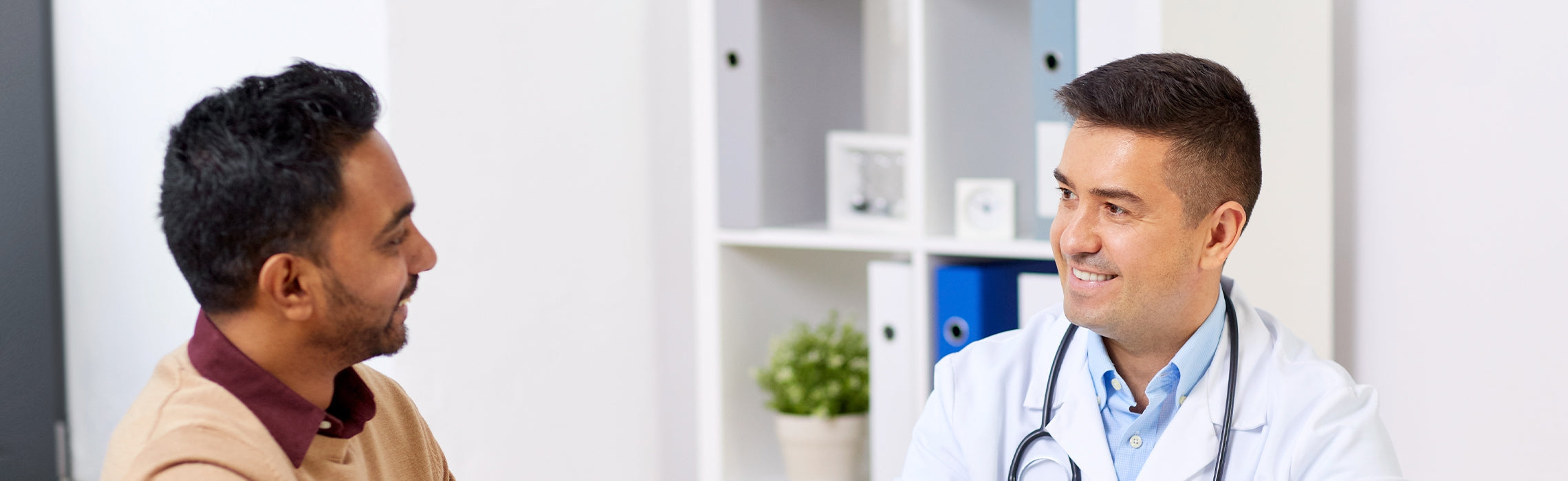 Healthcare professional wearing a lab coat and stethoscope sitting across from a patient in a medical office, engaging in conversation with shelves and binders in the background, representing doctor consultation, patient care, and healthcare services.