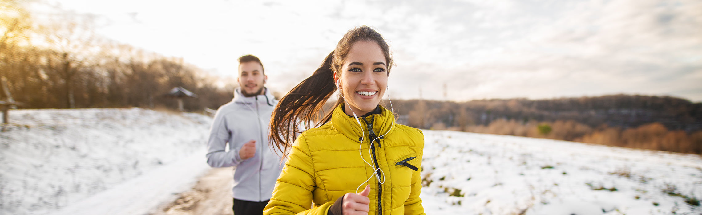 Two people jogging along a snowy outdoor trail, wearing winter jackets and earphones, with trees and open landscape in the background, representing cold‑weather running, fitness, and an active lifestyle.