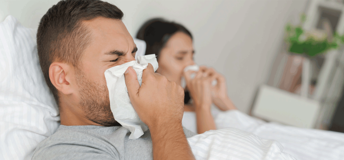 Two people sitting in bed under white blankets each hold tissues to their noses, suggesting cold or flu symptoms in a home setting. No visible text or branding.