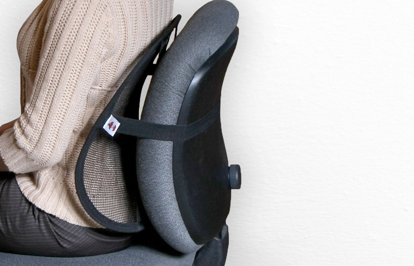 Close-up of a person seated in an office chair using a black ergonomic lumbar support cushion strapped to the chair back, promoting proper posture. No visible text or identifiable brand.