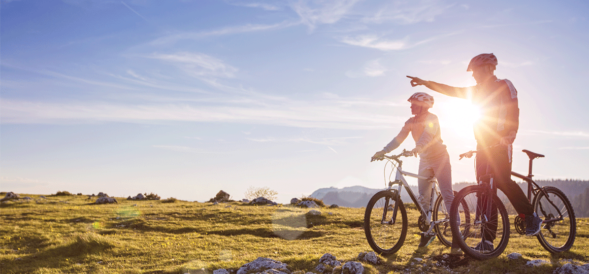 Two people with bicycles pause on a grassy hill at sunrise, one pointing into the distance as sunlight shines behind them in a scenic outdoor setting. No visible text or branding.