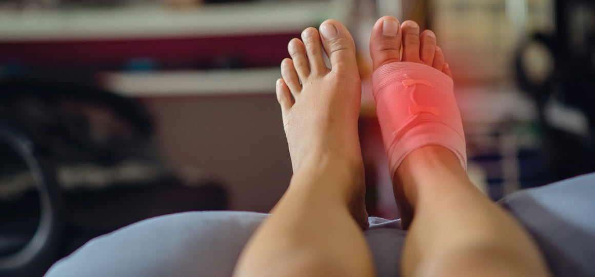Close-up of two feet resting on a bed, with one foot wrapped in a pink elastic bandage around the ankle, highlighted with a red glow to indicate pain or injury in a home setting. No visible text or branding.