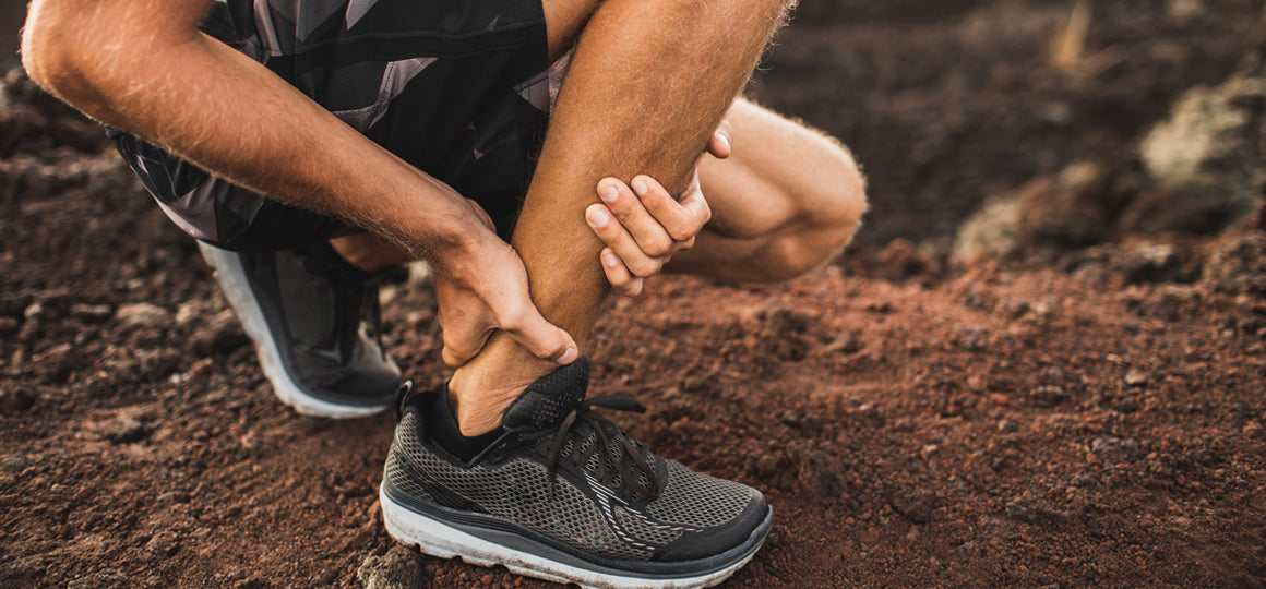 Close-up view of a person outdoors holding the lower part of their leg near the ankle. The individual is in a crouched position on uneven dirt ground, wearing black sneakers.