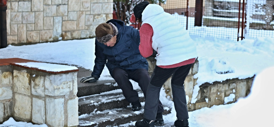 Person assisting another individual who has slipped on snowy outdoor steps, helping them regain balance near a stone wall in a winter setting. No visible text or branding.