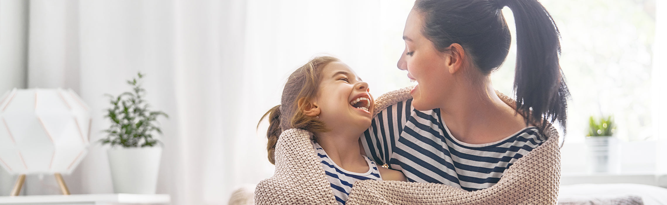 Adult and child wearing matching striped tops share a close embrace and look at each other in a bright living room, capturing a warm family moment. No visible text or branding.