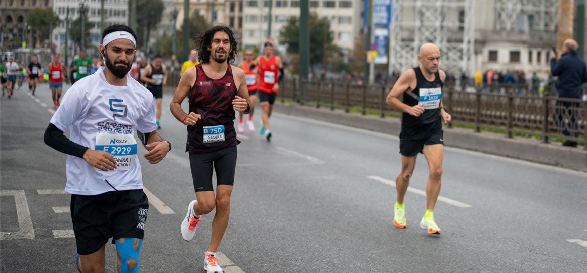 Runners participating in a road race on a city street wearing bib numbers and athletic gear, with buildings and spectators in the background. Visible text includes race bib numbers such as “E 2929” and “B 750.”