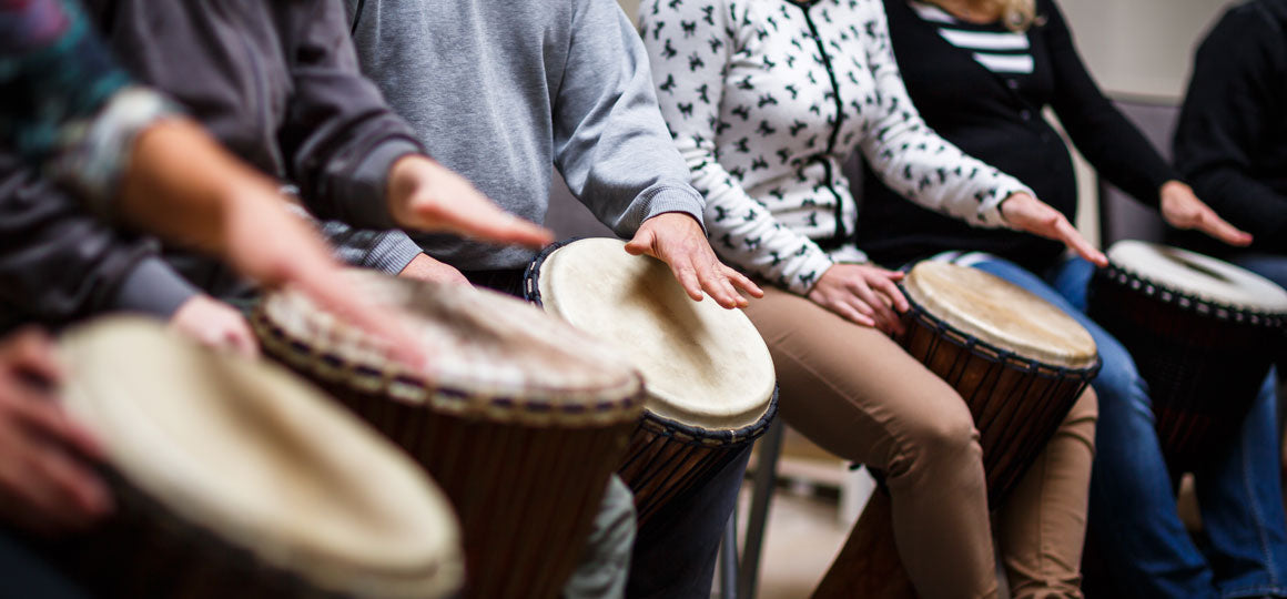 Group seated in a circle playing hand drums, each person tapping a djembe in an indoor group activity. No visible text or branding.