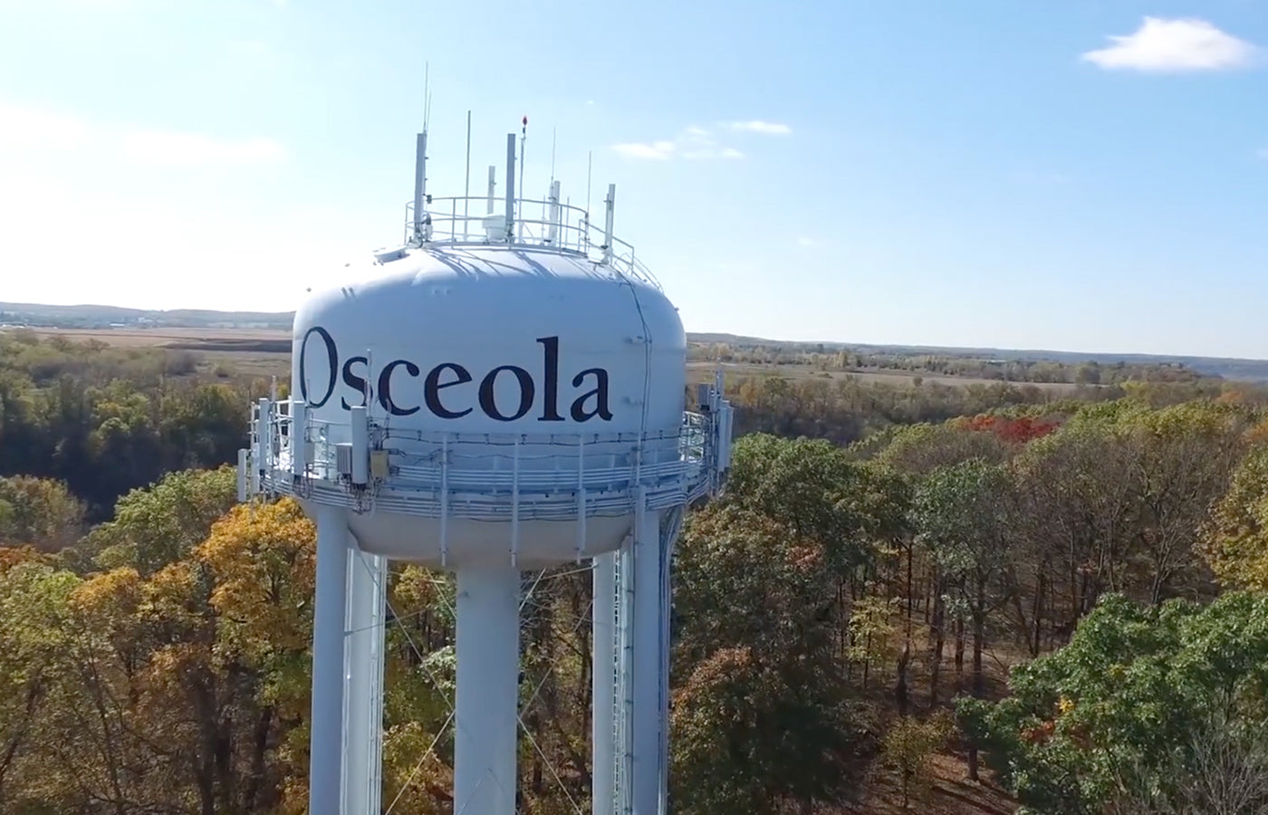White water tower with the visible text “Osceola” standing above a tree-filled landscape with autumn foliage under a clear blue sky, representing local infrastructure and a small-town setting.