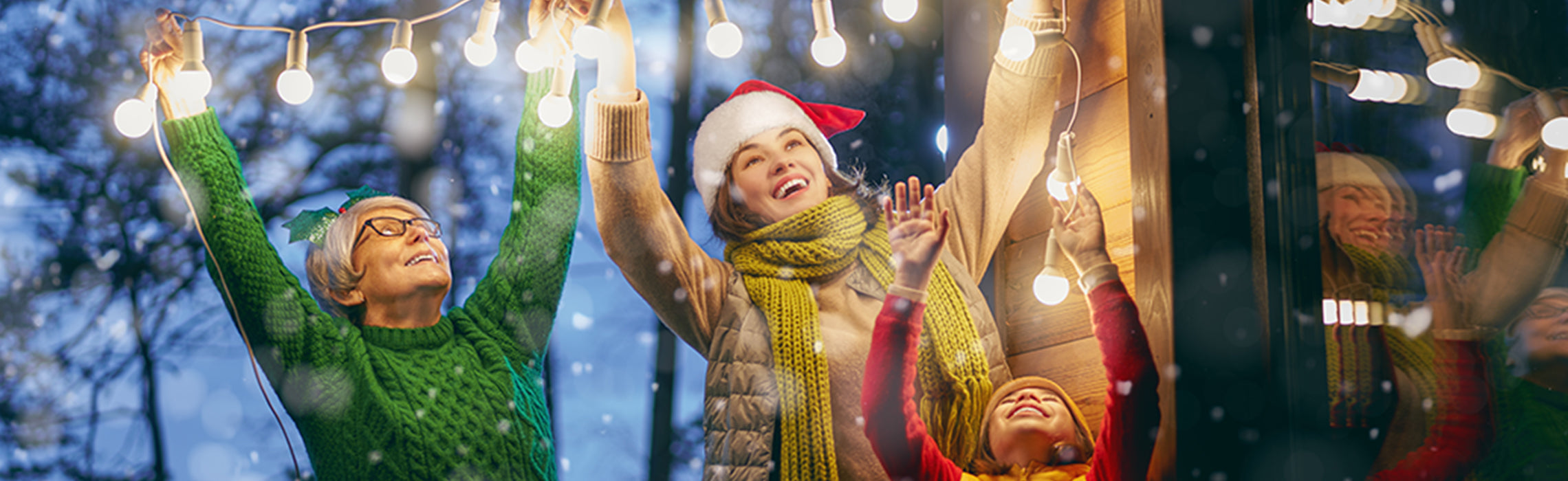 Three people in winter clothing and Santa hats hang string lights outdoors near a cabin as snow falls, creating a festive holiday scene. No visible text or branding.
