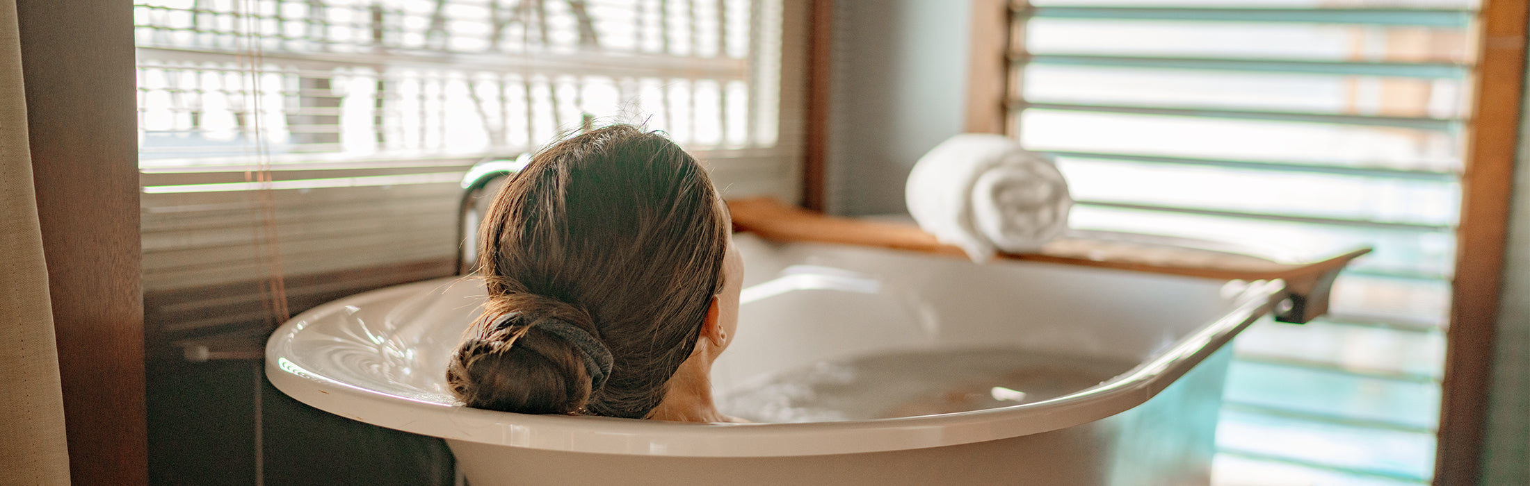 Person relaxing in a freestanding bathtub filled with water in a sunlit bathroom, with wooden accents, window blinds, and a rolled white towel nearby, suggesting a spa-like home setting. No visible text or branding.