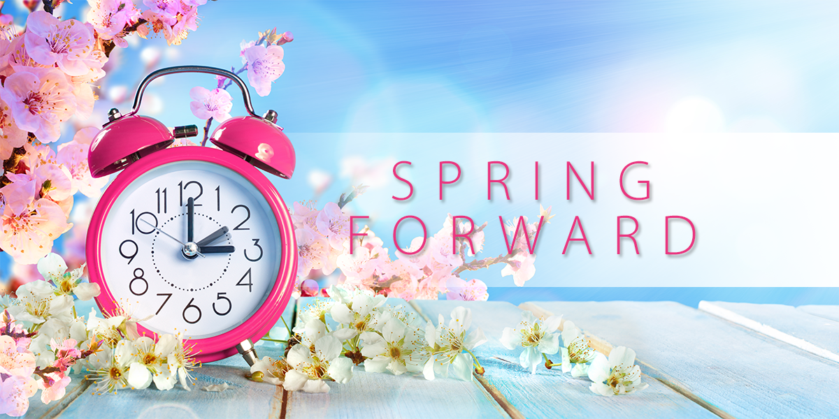 Pink alarm clock surrounded by spring blossoms on a wooden surface with a bright blue sky background and the text “SPRING FORWARD,” symbolizing daylight saving time.