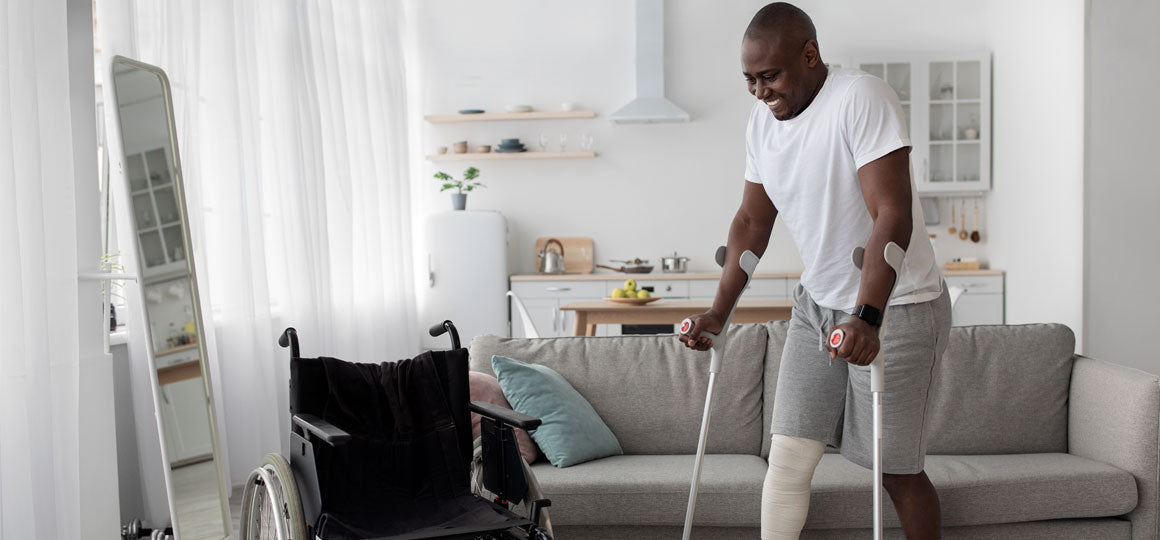 Person using crutches in a bright living room next to a wheelchair and couch, with a visible knee injury support, highlighting mobility recovery and at-home rehabilitation. No visible text or branding.